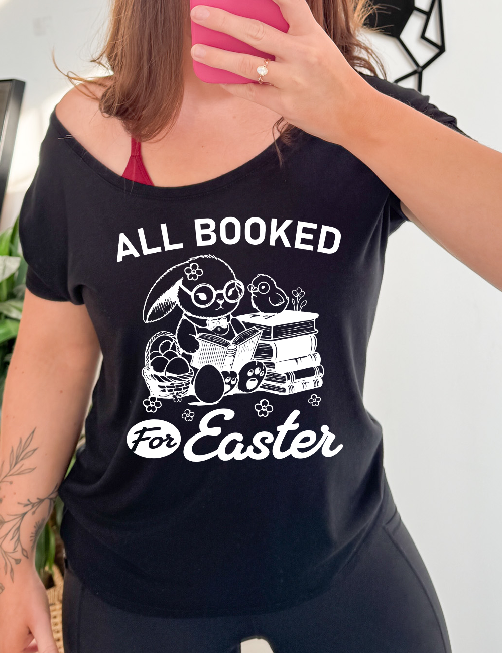 black slouchy tee with the words "all booked for Easter" on it, and the graphic of a bunny wearing glasses and a bowtie, reading a book, next to a stack of books (with a bird wearing glasses on top of it) on one side, and a basket of eggs on the other side