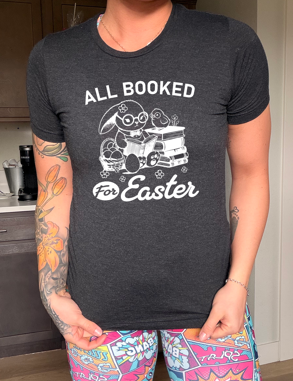 black unisex tee with the words "all booked for Easter" on it, and the graphic of a bunny wearing glasses and a bowtie, reading a book, next to a stack of books (with a bird wearing glasses on top of it) on one side, and a basket of eggs on the other side