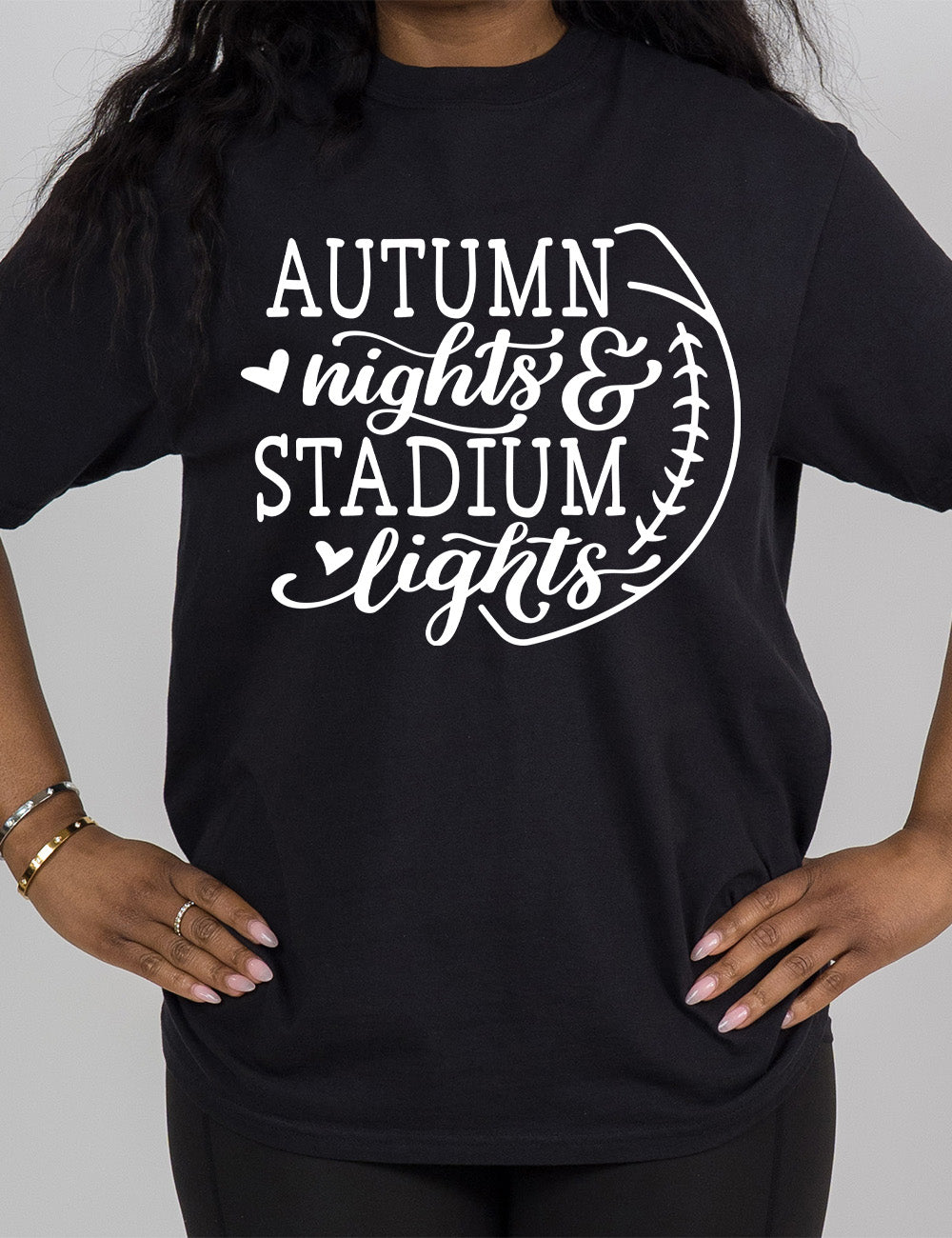 black modern tee with the words "autumn nights & stadium lights" in both regular and cursive font, slightly overlapping the outline of a football, and with a little heart in front of "nights" and "lights"