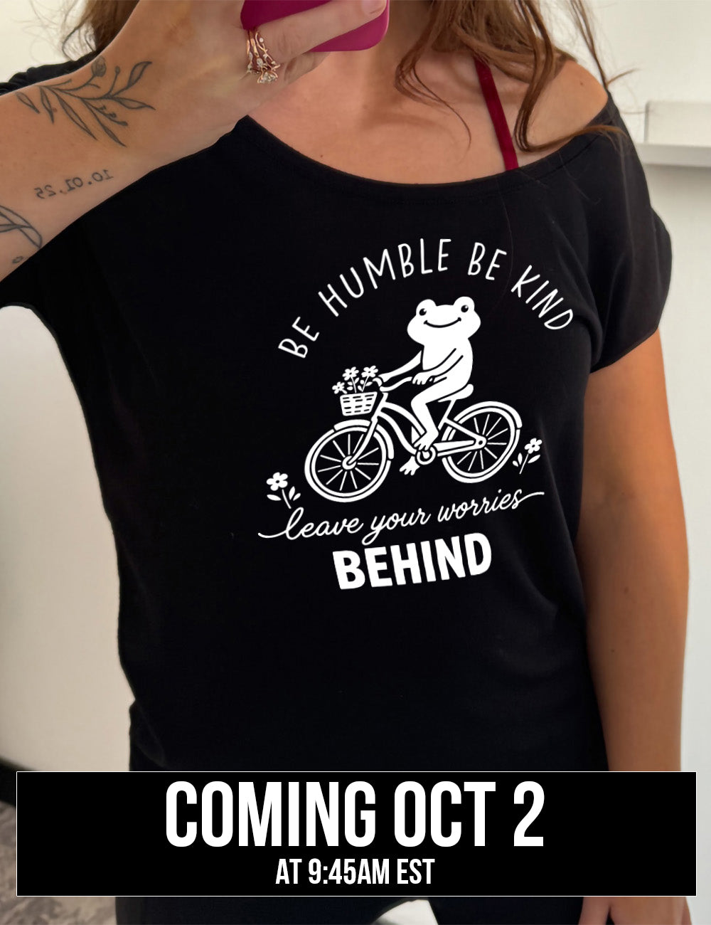 black slouchy tee with the graphic of a happy frog riding a bike that has a basket of flowers attached, with a flower in front and behind the bike, and the wording arching above and ending below saying "be humble be kind leave your worries BEHIND", coming October 2nd