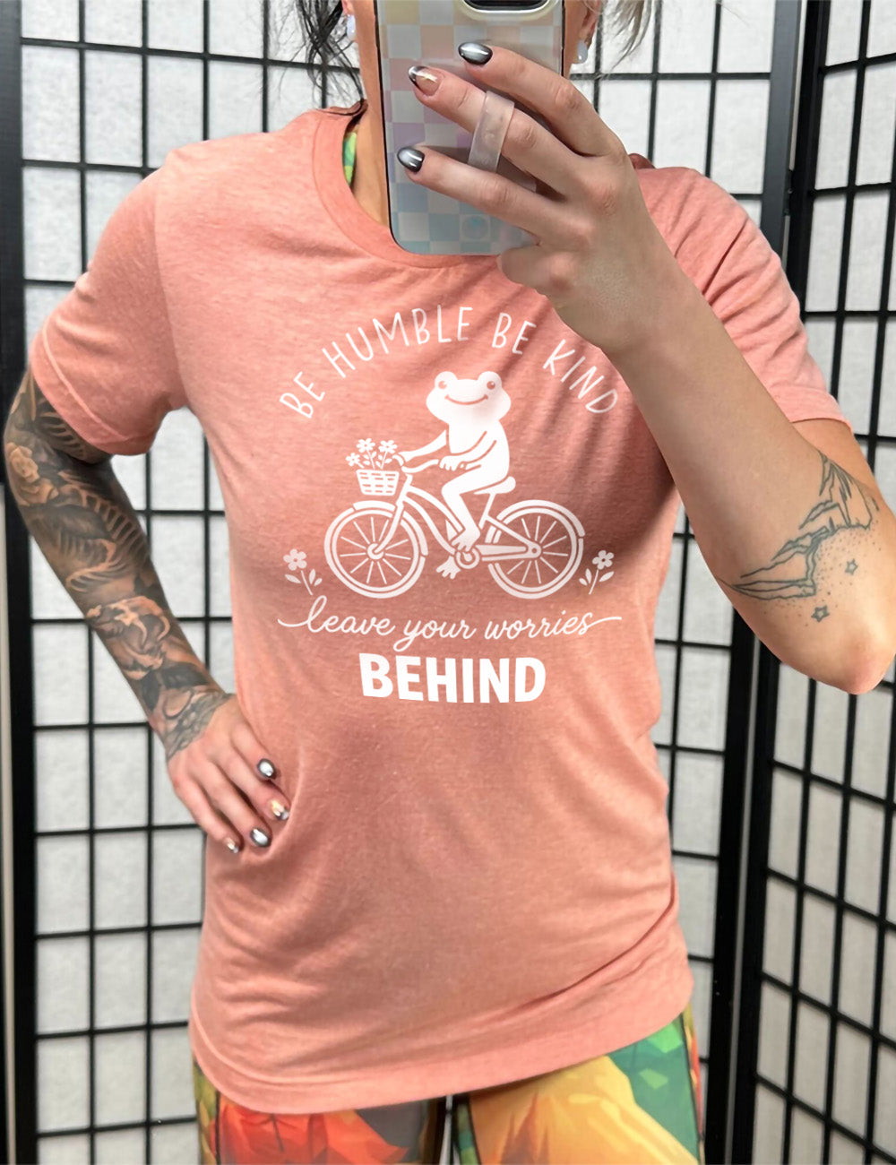sunset unisex tee with the graphic of a happy frog riding a bike that has a basket of flowers attached, with a flower in front and behind the bike, and the wording arching above and ending below saying "be humble be kind leave your worries BEHIND"