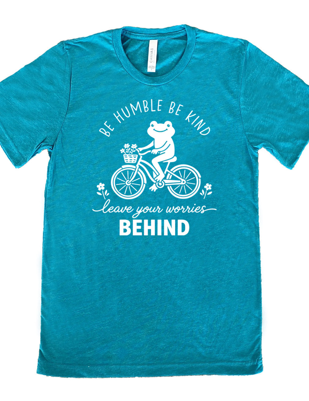 teal unisex tee with the graphic of a happy frog riding a bike that has a basket of flowers attached, with a flower in front and behind the bike, and the wording arching above and ending below saying "be humble be kind leave your worries BEHIND"