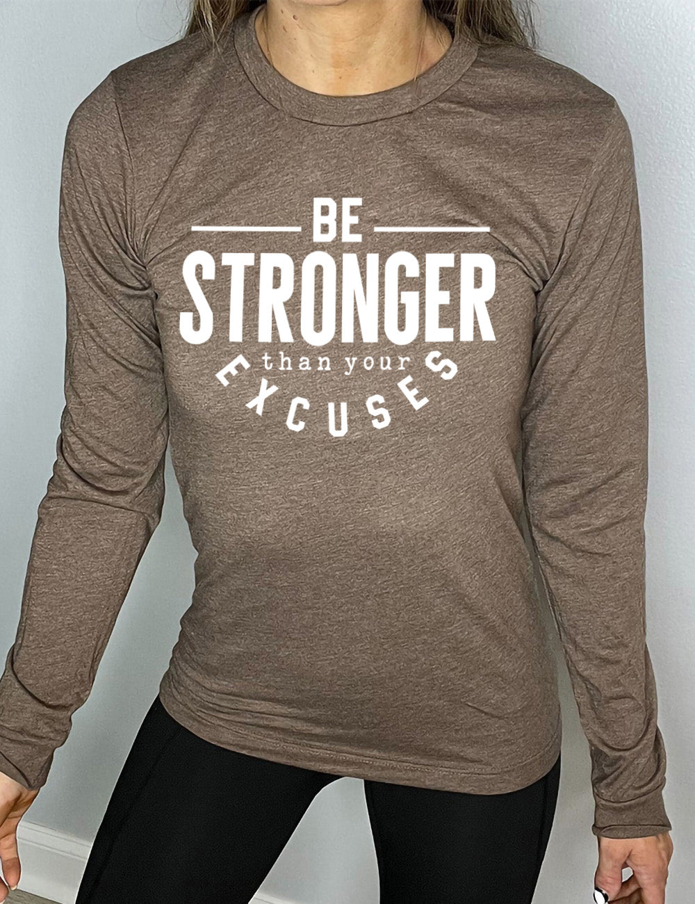 heather brown long sleeve tee with words on it saying "be STRONGER than your excuses", with lines in front and after the word "be", the word "stronger" in the largest font, and the word "excuses" arching 
