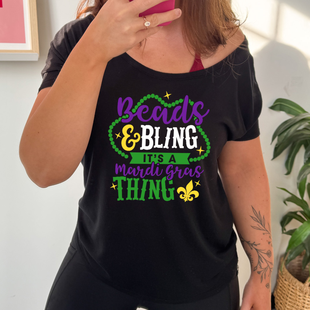 black slouchy tee with the words "beads & bling it's a mardi gras thing" on it, with graphics of a ring of beads around part of the words and a few stars as well as a fleur-de-lis symbol