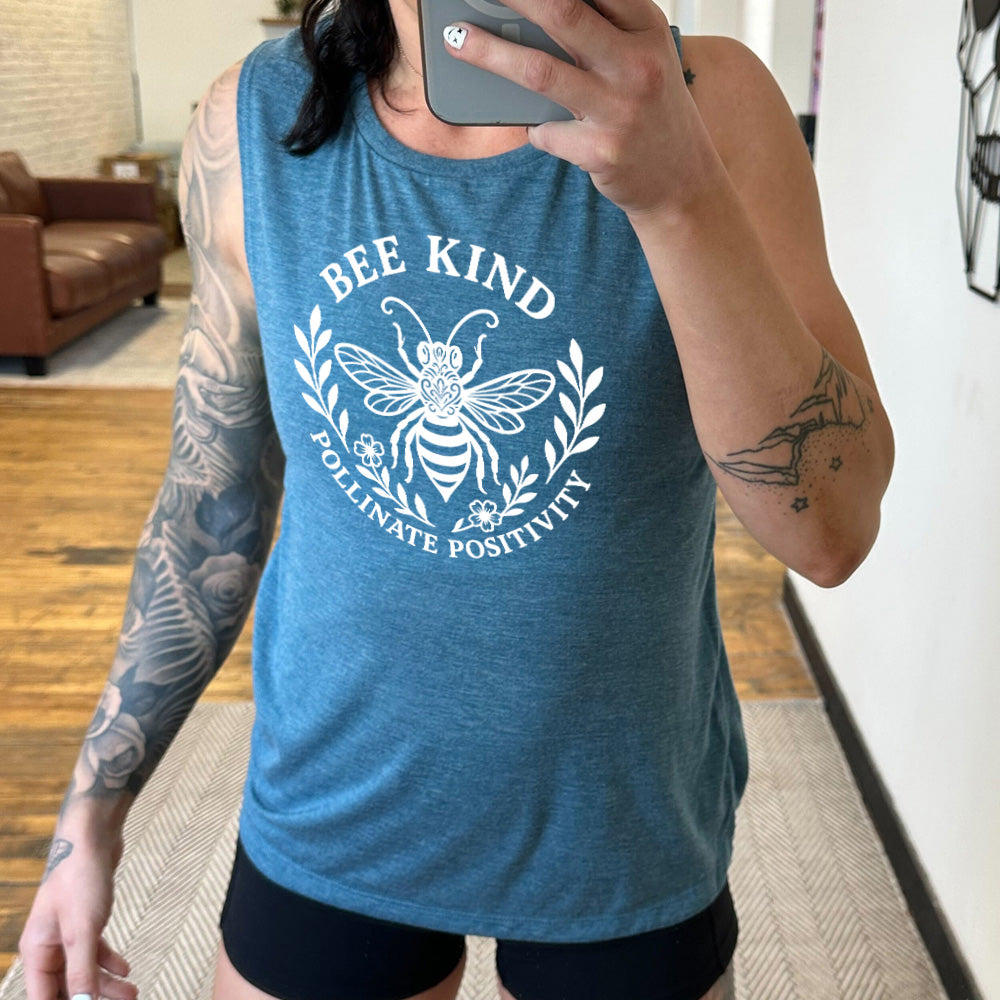 deep teal muscle tank with the words "bee kind pollinate positivity" on it, arranged in two arcs, forming a circled effect, with two branches and two flowered branches within the circle, and a large fancy bee in the center