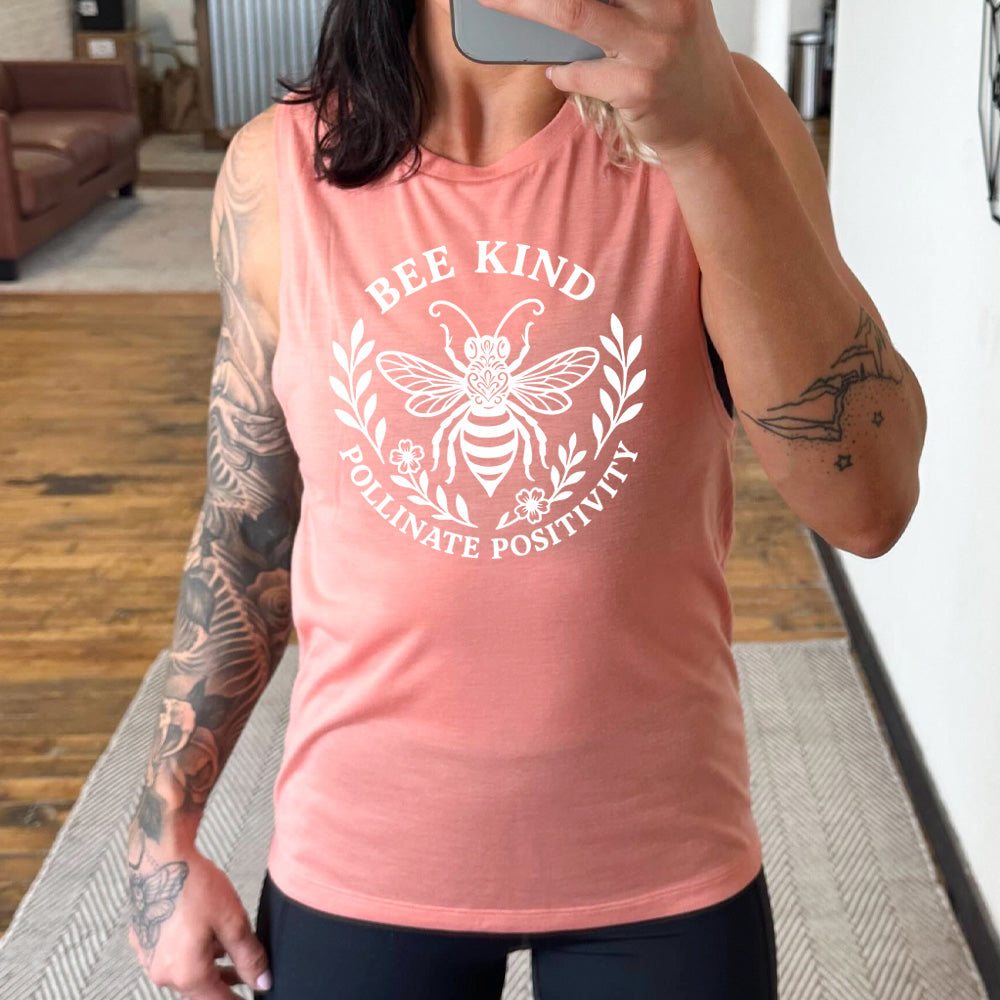 peach muscle tank with the words "bee kind pollinate positivity" on it, arranged in two arcs, forming a circled effect, with two branches and two flowered branches within the circle, and a large fancy bee in the center
