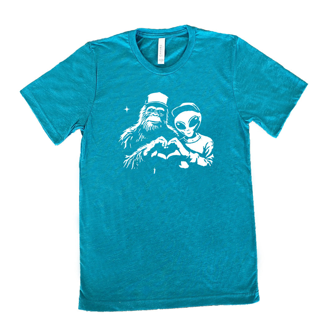 Teal colored shirt with bigfoot and alien doing heart hands graphic on it