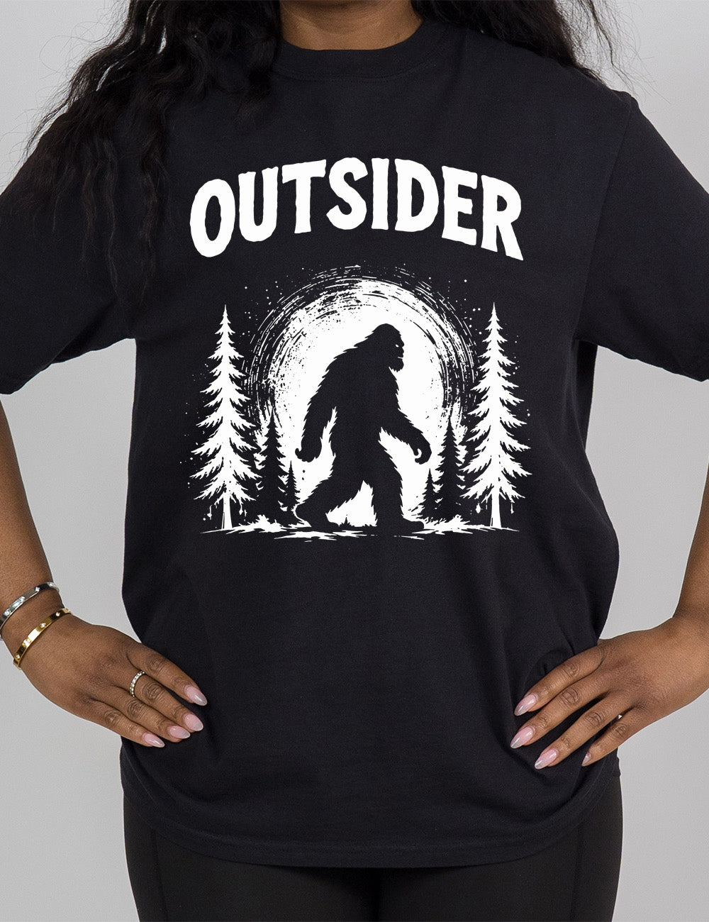 black modern tee with the graphic of bigfoot, silhouetted against a large moon, walking through pine trees, and a word above it saying "OUTSIDER"