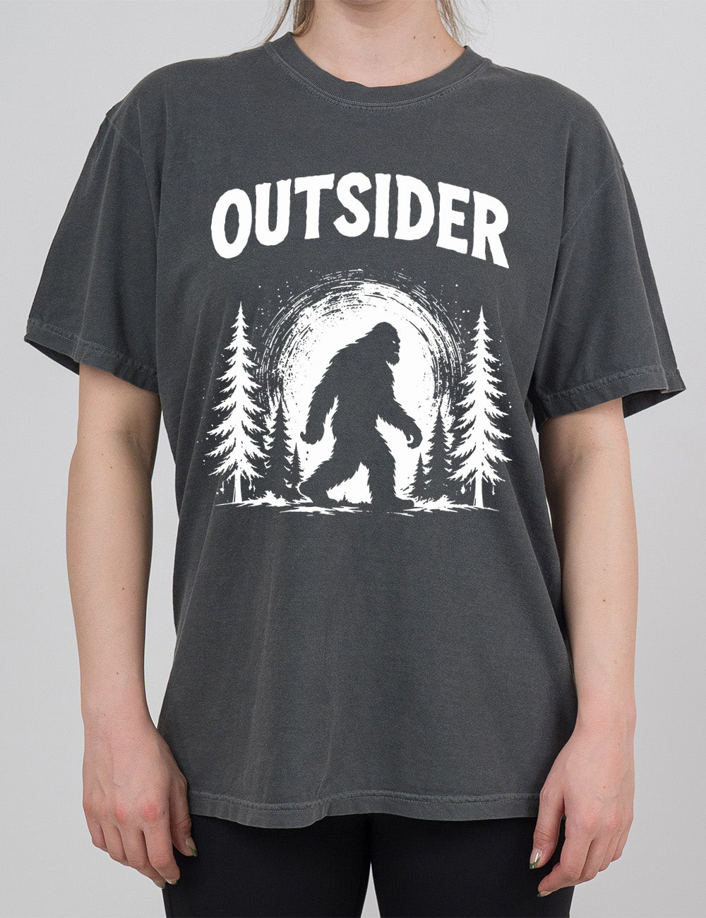 pepper modern tee with the graphic of bigfoot, silhouetted against a large moon, walking through pine trees, and a word above it saying "OUTSIDER"