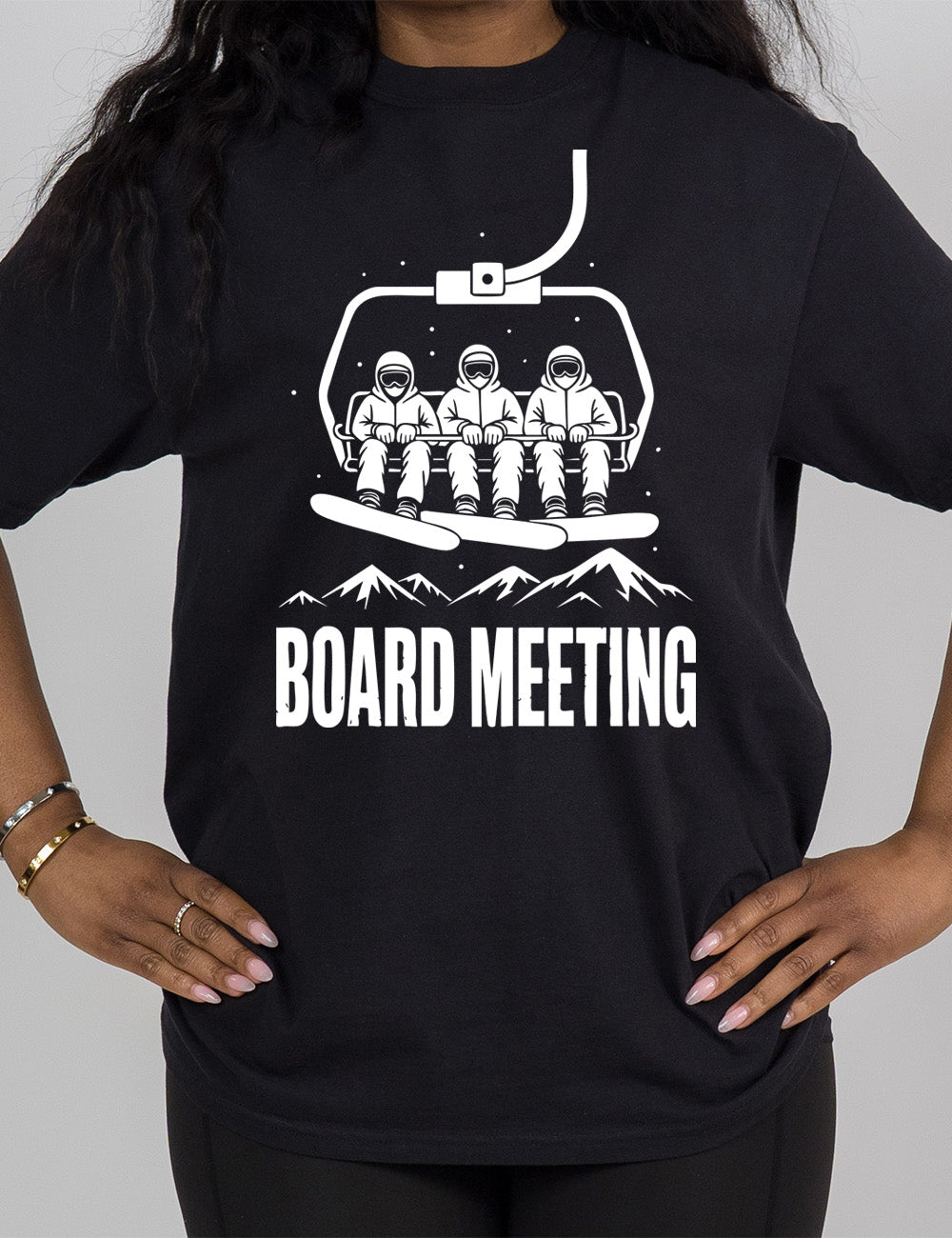 black modern tee with the graphic of three snowboarders riding a ski lift, with snowy mountains in the distance and little dots of snow around, and words underneath this saying "board meeting" in capital letters
