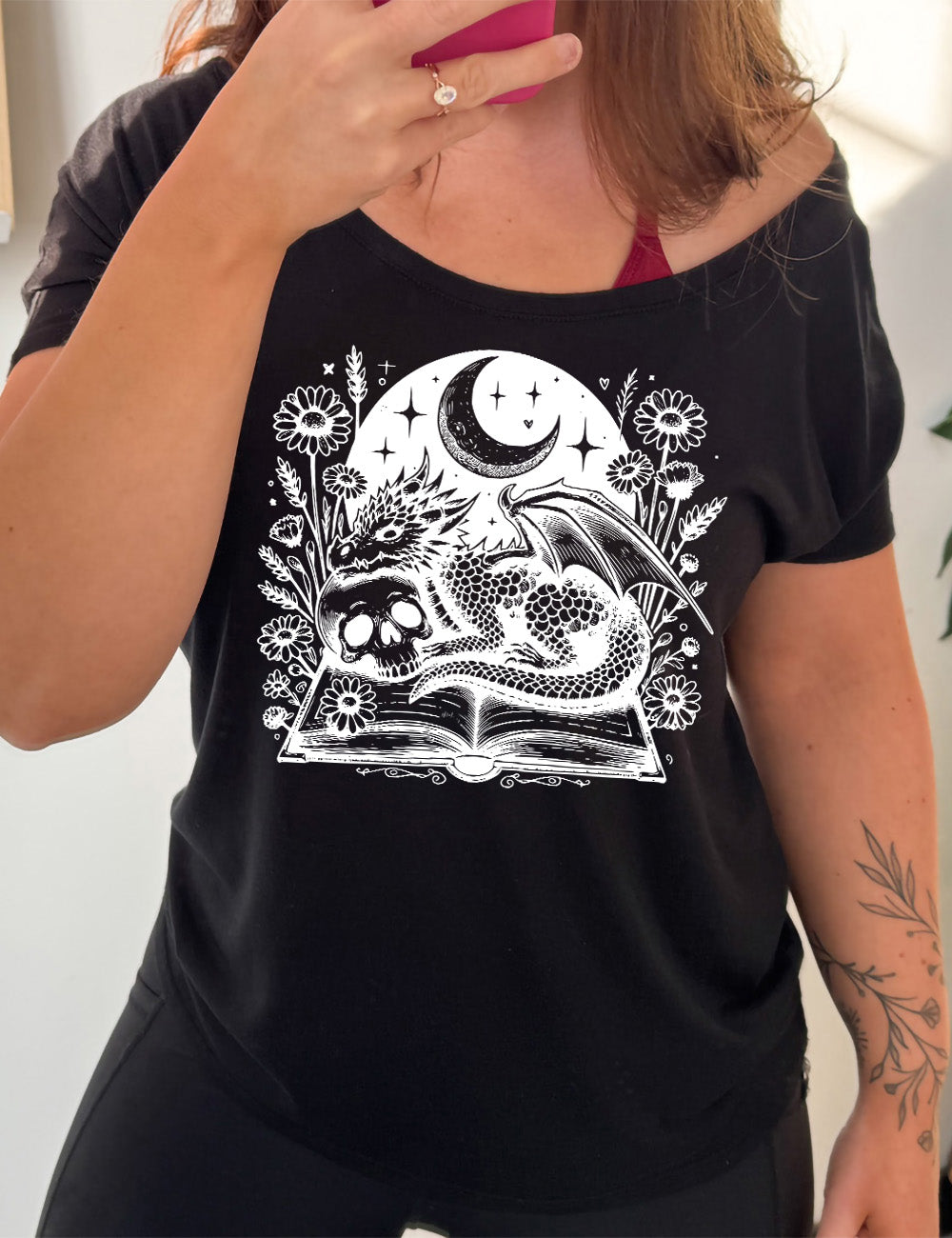 black slouchy tee with the very detailed and textured graphic of a dragon and a skull resting on an open book, with night sky behind it, with a large crescent moon, and many flowers growing around this scene