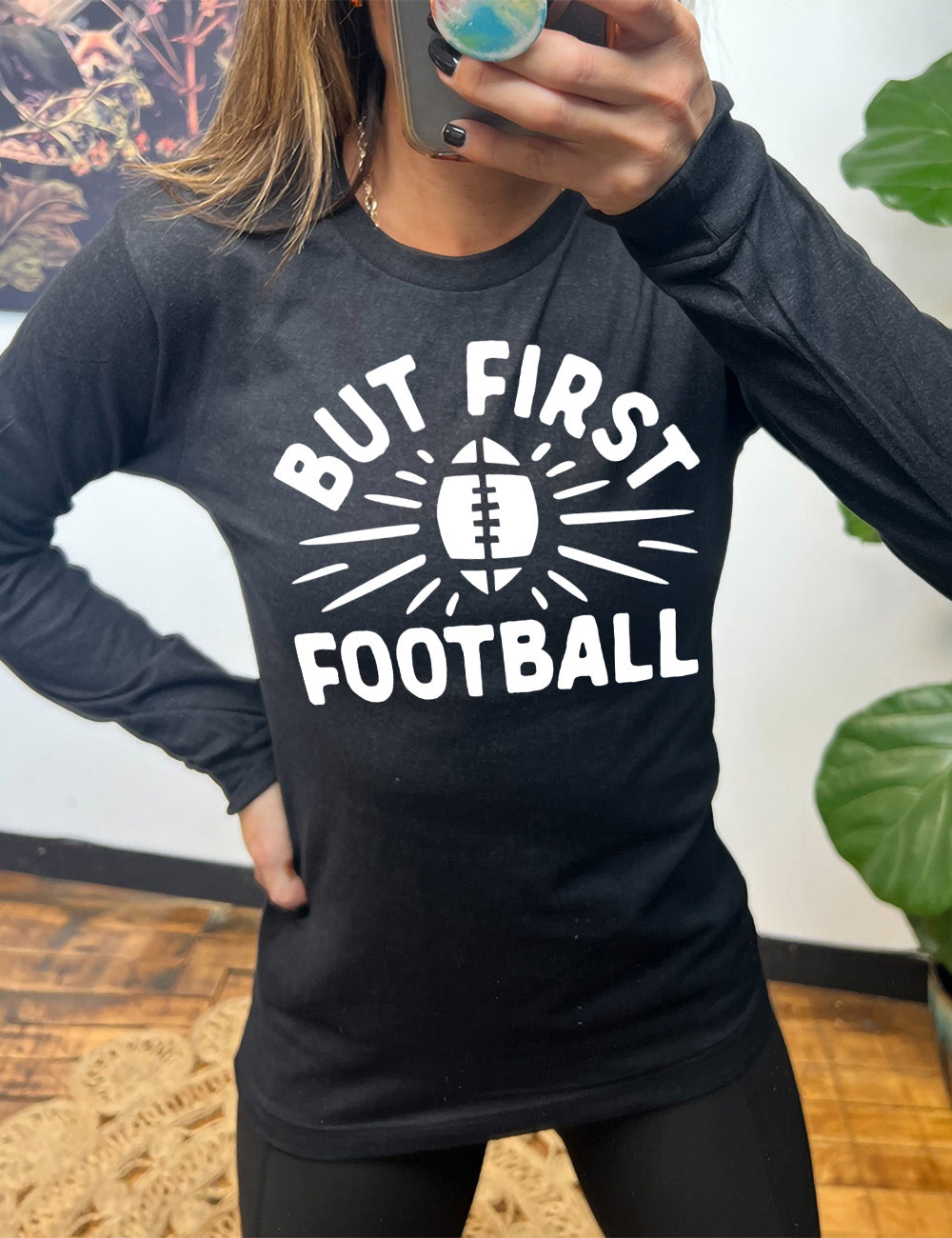 heather black long sleeve with the words "but first football" in capital font, with "But first" arching over the graphic of a football, with rays beaming off of it