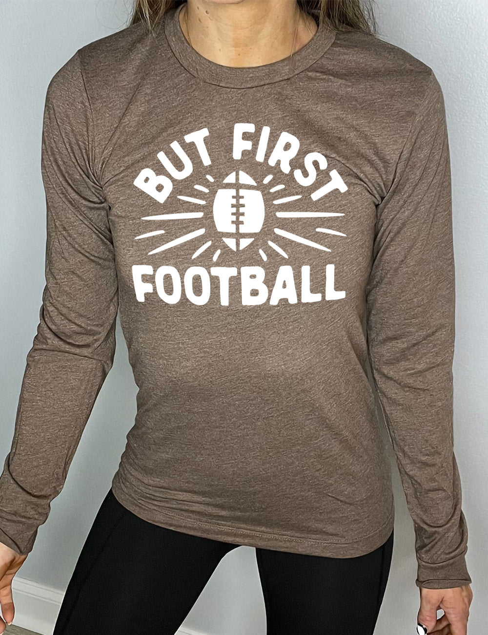 heather brown long sleeve with the words "but first football" in capital font, with "But first" arching over the graphic of a football, with rays beaming off of it