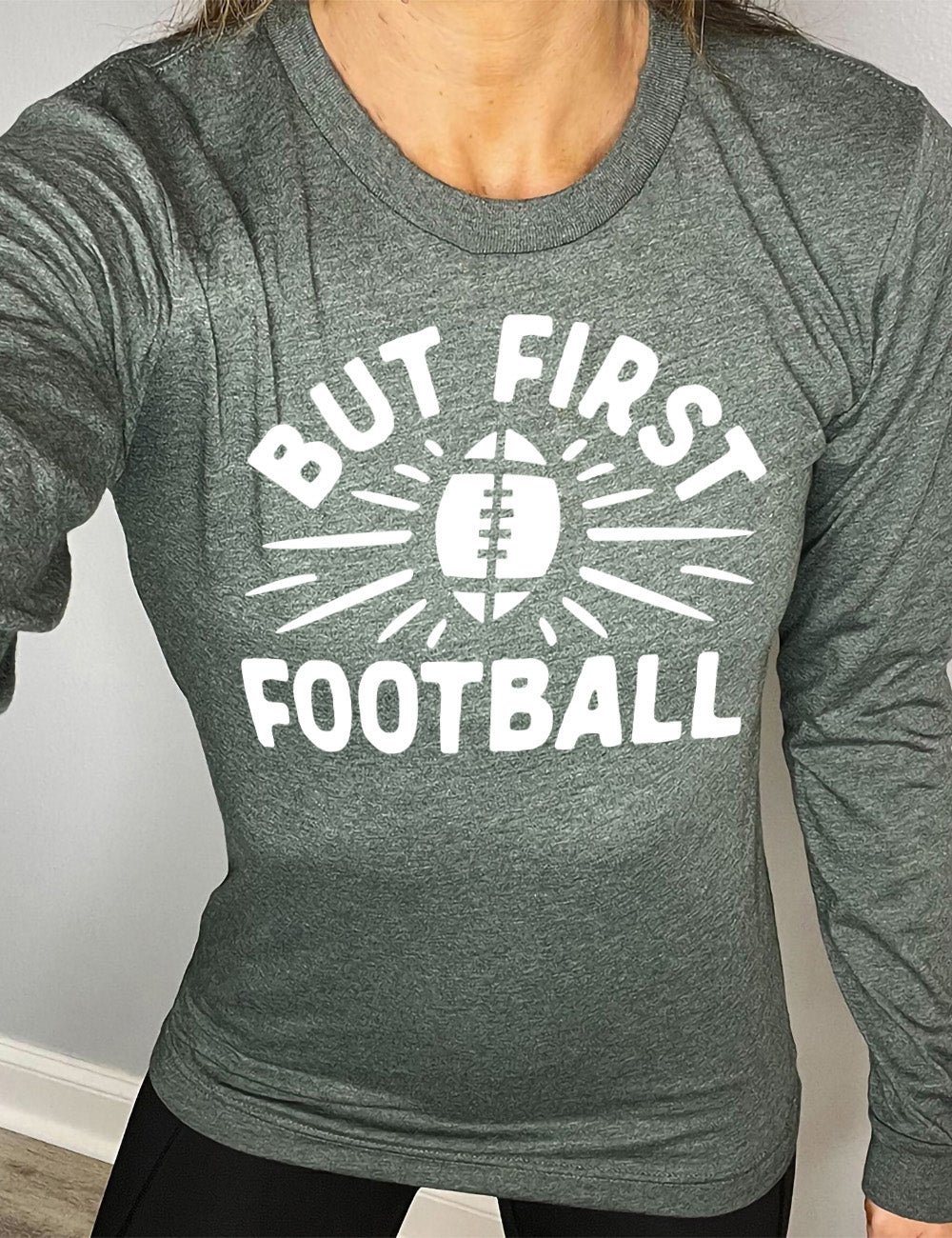 heather forest long sleeve with the words "but first football" in capital font, with "But first" arching over the graphic of a football, with rays beaming off of it