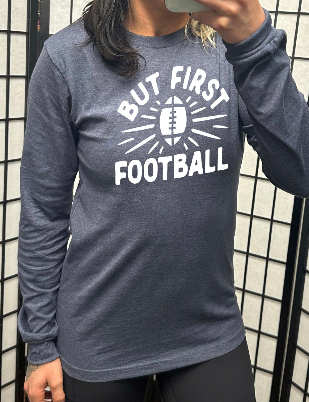 heather navy long sleeve with the words "but first football" in capital font, with "But first" arching over the graphic of a football, with rays beaming off of it