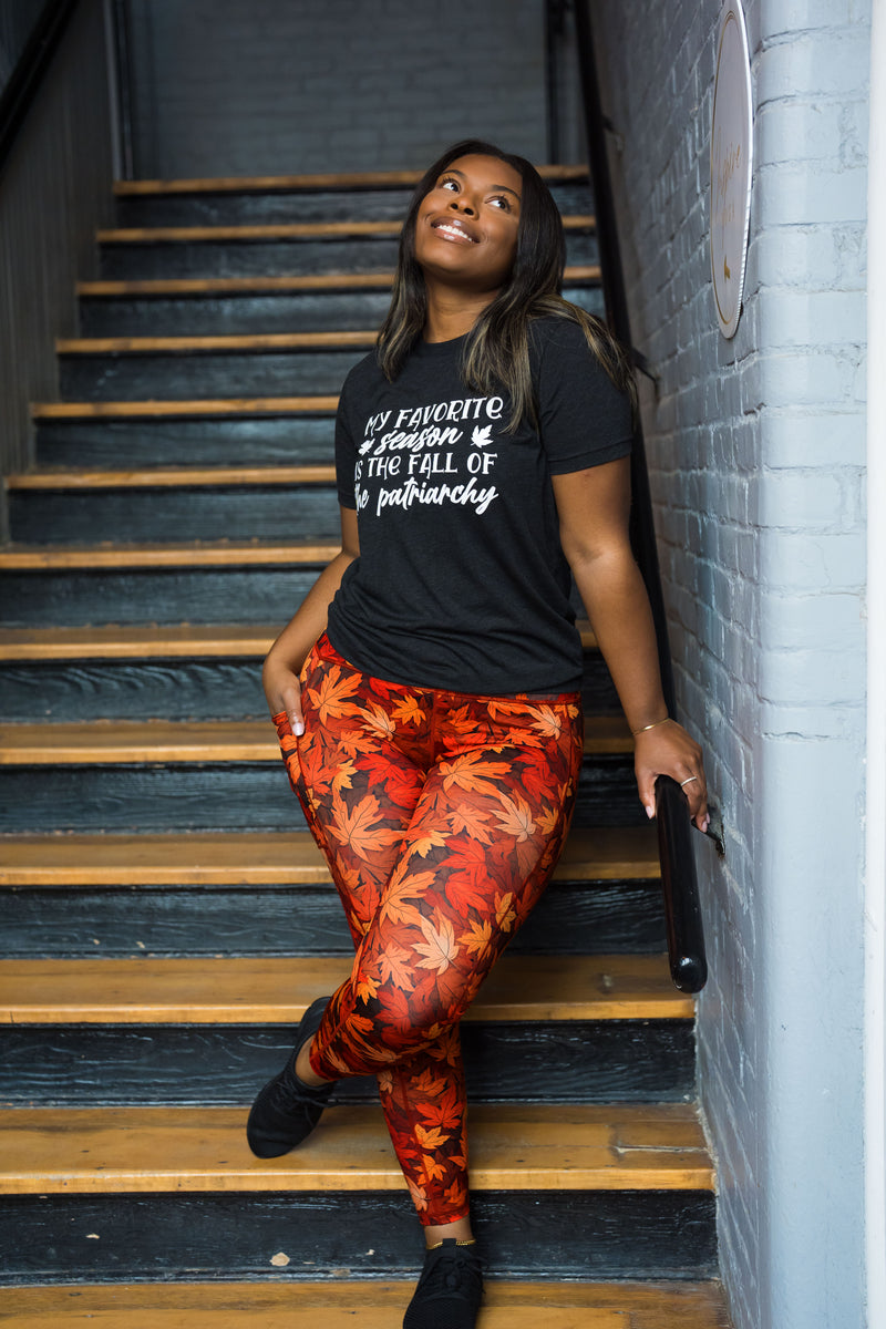 A woman posing on an indoor staircase wearing a black graphic t-shirt and red/orange autumn leaf print leggings.