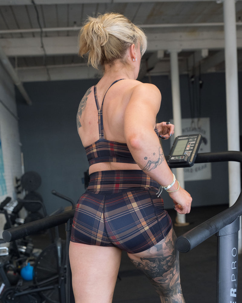 A woman with blonde hair in a ponytail, wearing a matching plaid athletic sports bra and shorts set, running on a treadmill in a gym.