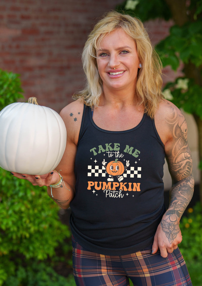 A smiling woman outdoors wearing a black tank top with a 'Take Me to the Pumpkin Patch' graphic and plaid pants, while holding a white pumpkin.