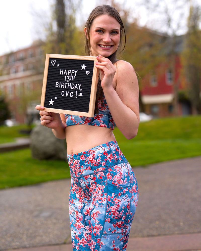 a model wearing cherry blossom print activewear holding a sign that says "Happy 13th Birthday CVG!"