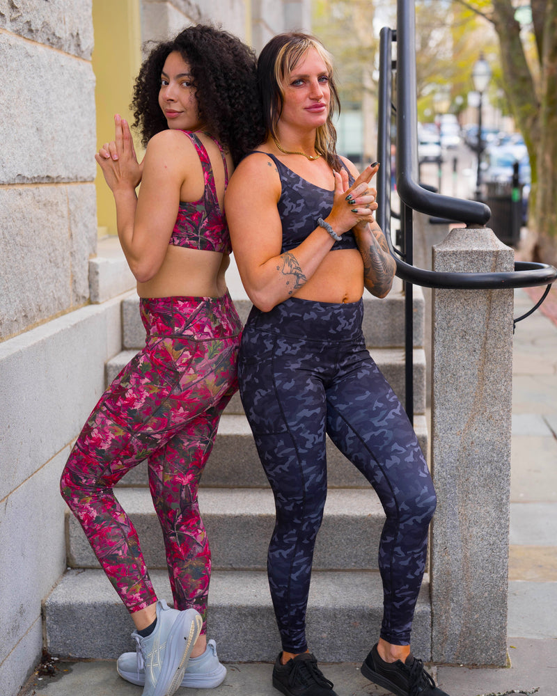 Two women posing together on outdoor stone steps wearing matching athletic sets. The woman on the left wears a pink camo sports bra and matching leggings, while the woman on the right wears a dark blue-gray camo patterned sports bra and matching leggings.