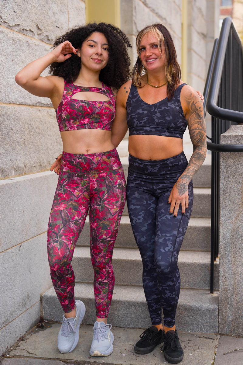 Two women posing on outdoor stone steps wearing matching athletic sets. The woman on the left wears a pink camo sports bra and leggings with white sneakers, while the woman on the right wears a dark camouflage sports bra and matching leggings with black sneakers.