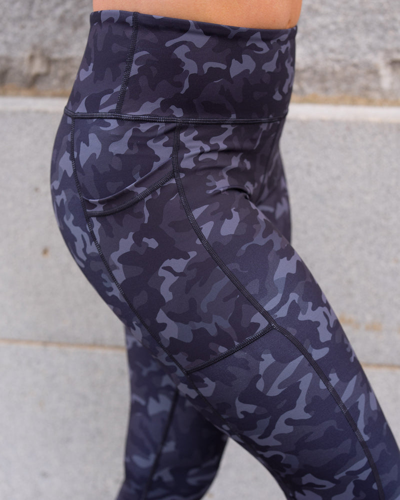 A close-up side view of a person wearing high-waisted leggings with a dark blue and gray camouflage pattern, standing against a concrete wall.