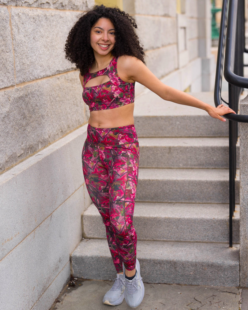 A woman with curly dark hair stands on outdoor stone steps wearing a matching camo print activewear set consisting of a sports bra and leggings in a vibrant pink color paired with light gray sneakers