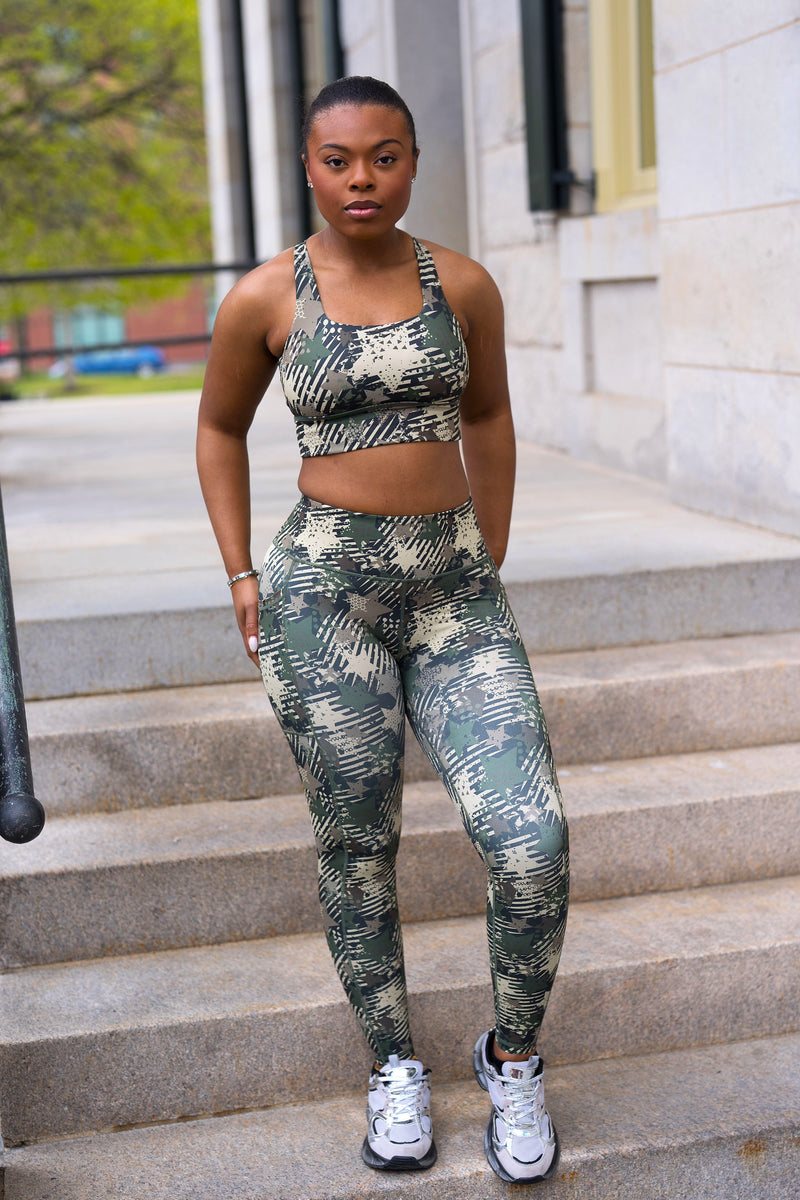 A woman stands on outdoor stone steps wearing a matching camouflage print sports bra and leggings set with white sneakers.
