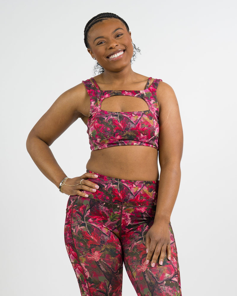 A woman modeling a matching activewear set featuring a pink camouflage print. The set includes a sports bra and matching high-waisted leggings, photographed against a solid light gray background.