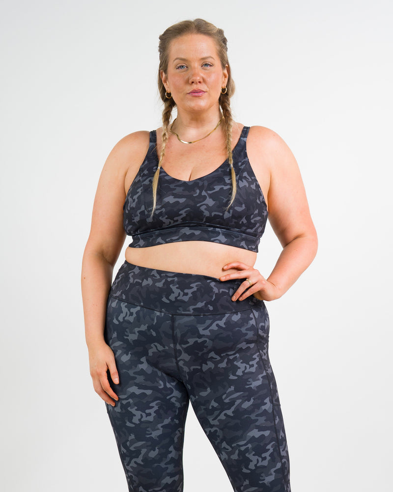 A woman modeling a matching camouflage print activewear set, consisting of a sports bra and high-waisted leggings, against a plain white studio background.