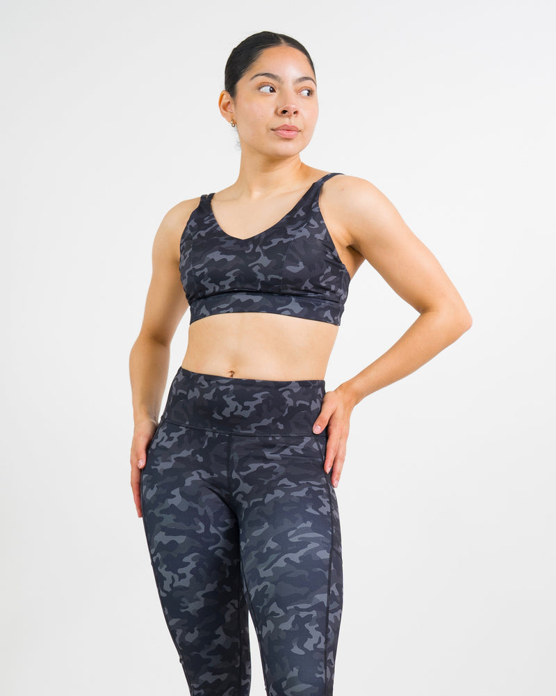 A woman modeling a matching camouflage print activewear set, consisting of a sports bra and high-waisted leggings, against a plain white studio background.