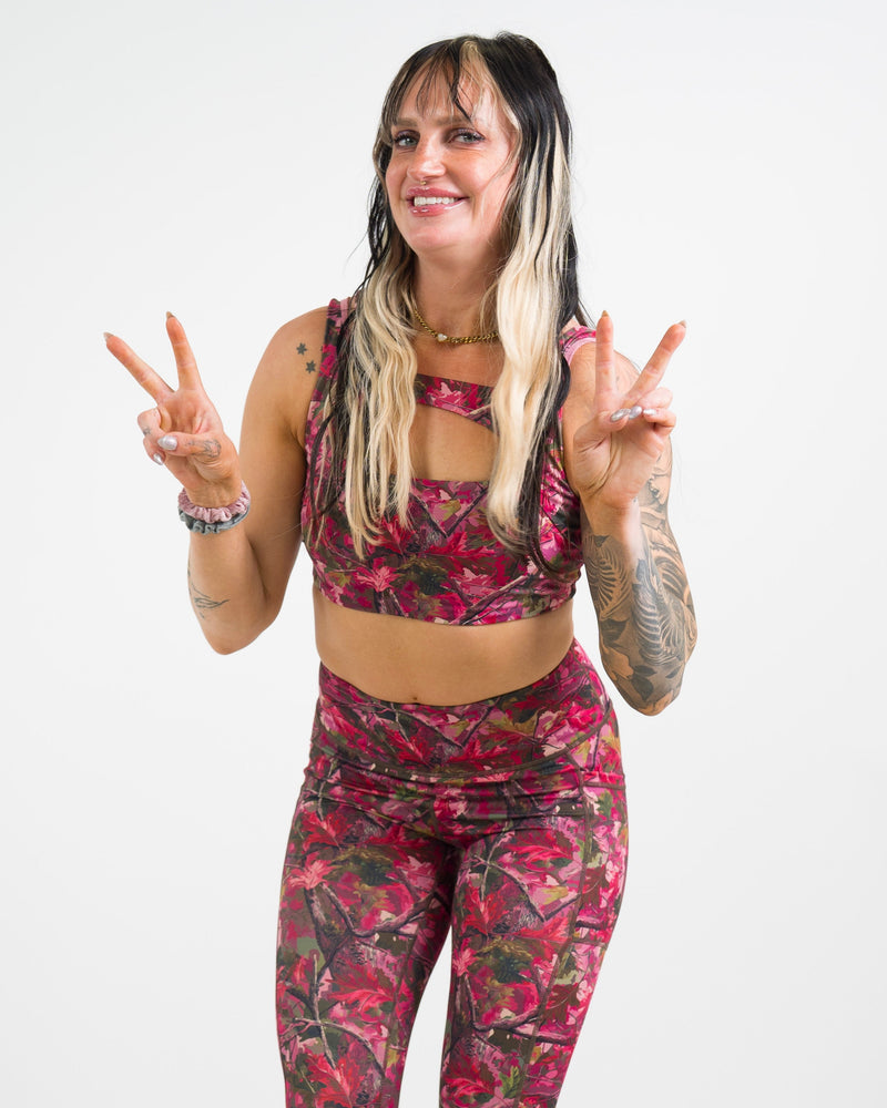 A woman modeling a pink camo print activewear set, posing with peace signs against a white studio background.