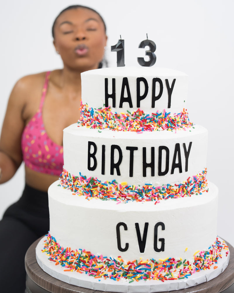 A three-tier white birthday cake with black text reading 'HAPPY BIRTHDAY' and '13' candles on top, decorated with colorful sprinkles. A woman in a pink patterned sports bra is blurred in the background blowing a kiss.