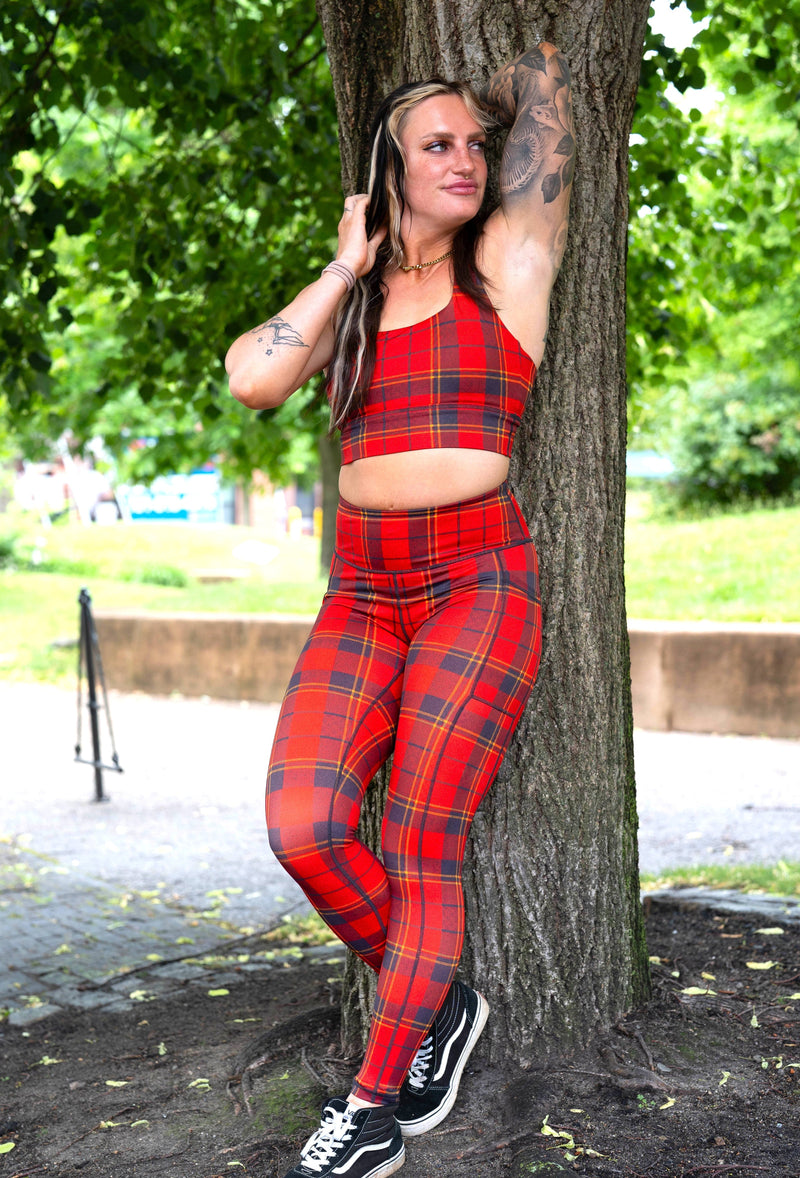 A woman wearing a matching red and black plaid activewear set, leaning against a large tree in an outdoor park setting.