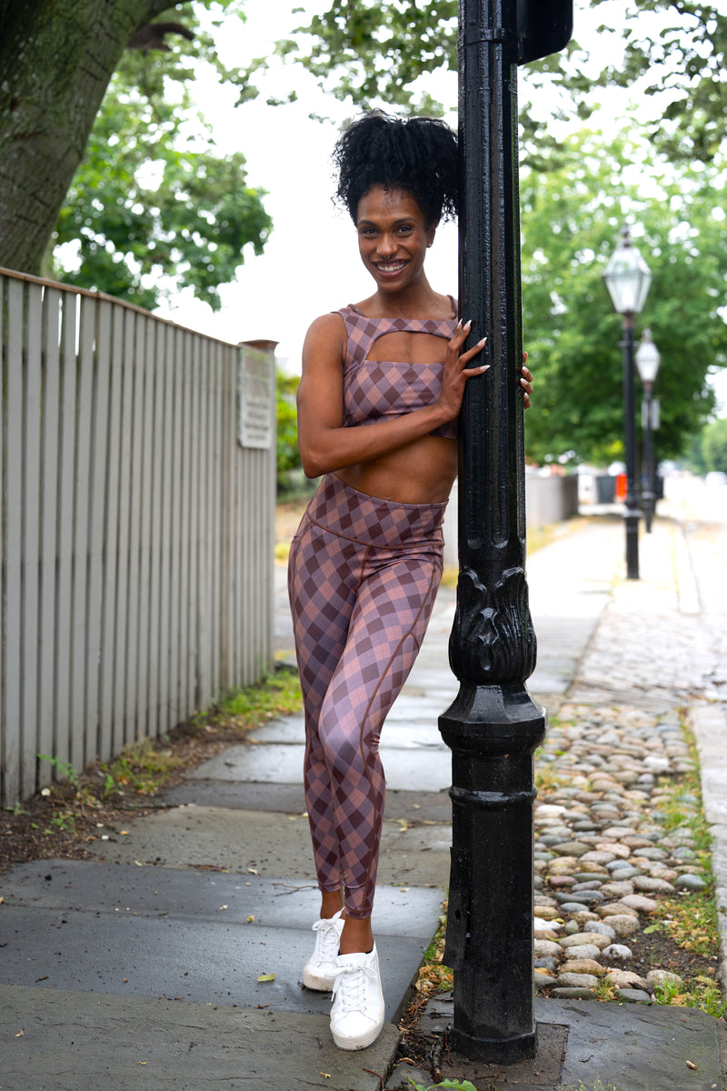 A woman posing outdoors on a sidewalk next to a black lamppost, wearing a matching two-piece athletic set with a dusty rose and purple plaid pattern, white sneakers, and leaning against the post.