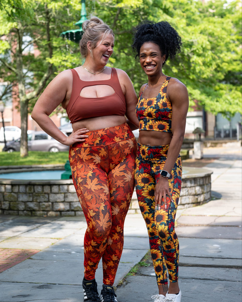 Two women standing outdoors on a paved walkway in a park-like setting, wearing matching activewear sets. 