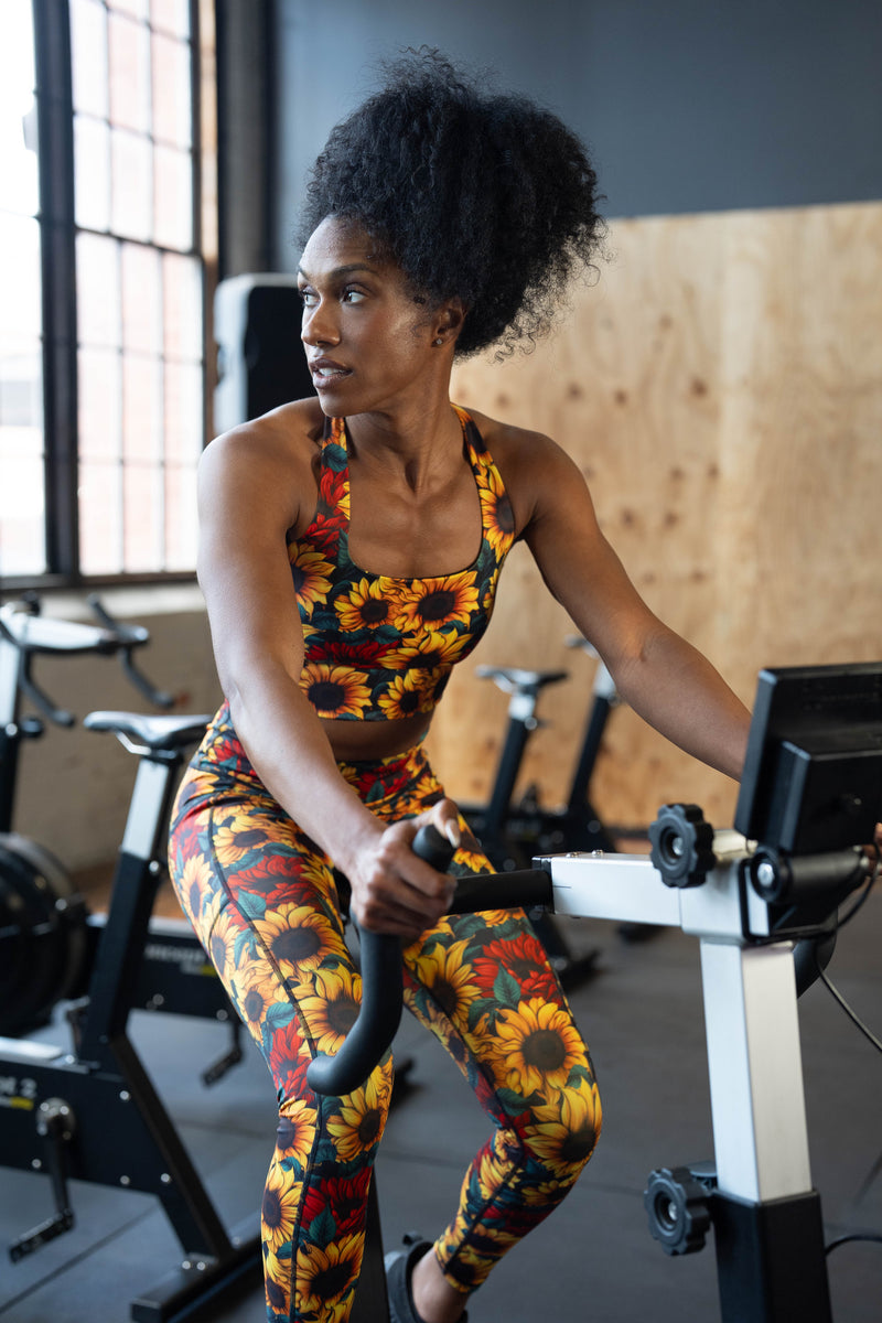 A woman with dark curly hair exercises on a stationary bike in a gym, wearing a matching floral print sports bra and leggings set with a sunflower pattern.