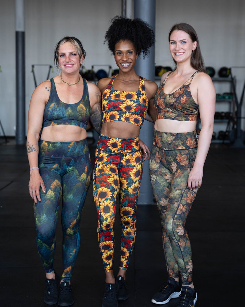 Three women standing together in a gym setting, each wearing a matching sports bra and leggings activewear set with different colorful patterns: a blue and green abstract print, a yellow, orange, and red sunflower print, and a green and brown camouflage print.