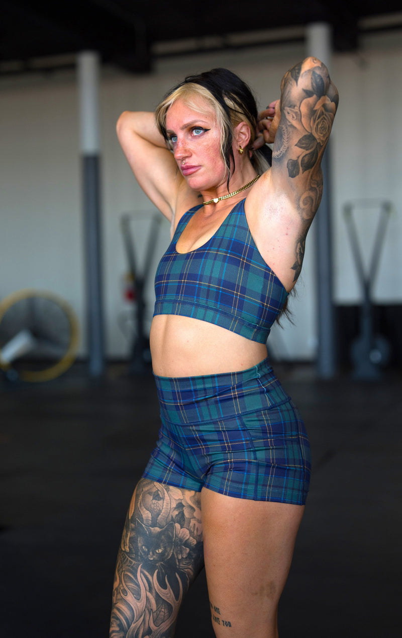 A woman with tattoos poses in a gym wearing a matching blue and green plaid sports bra and shorts set.