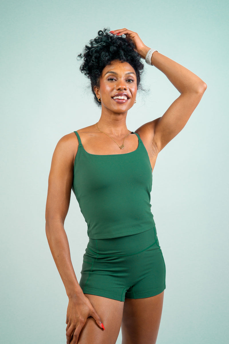A woman modeling a matching green two-piece activewear set consisting of a tank top and shorts, posing against a solid light blue-green studio background.