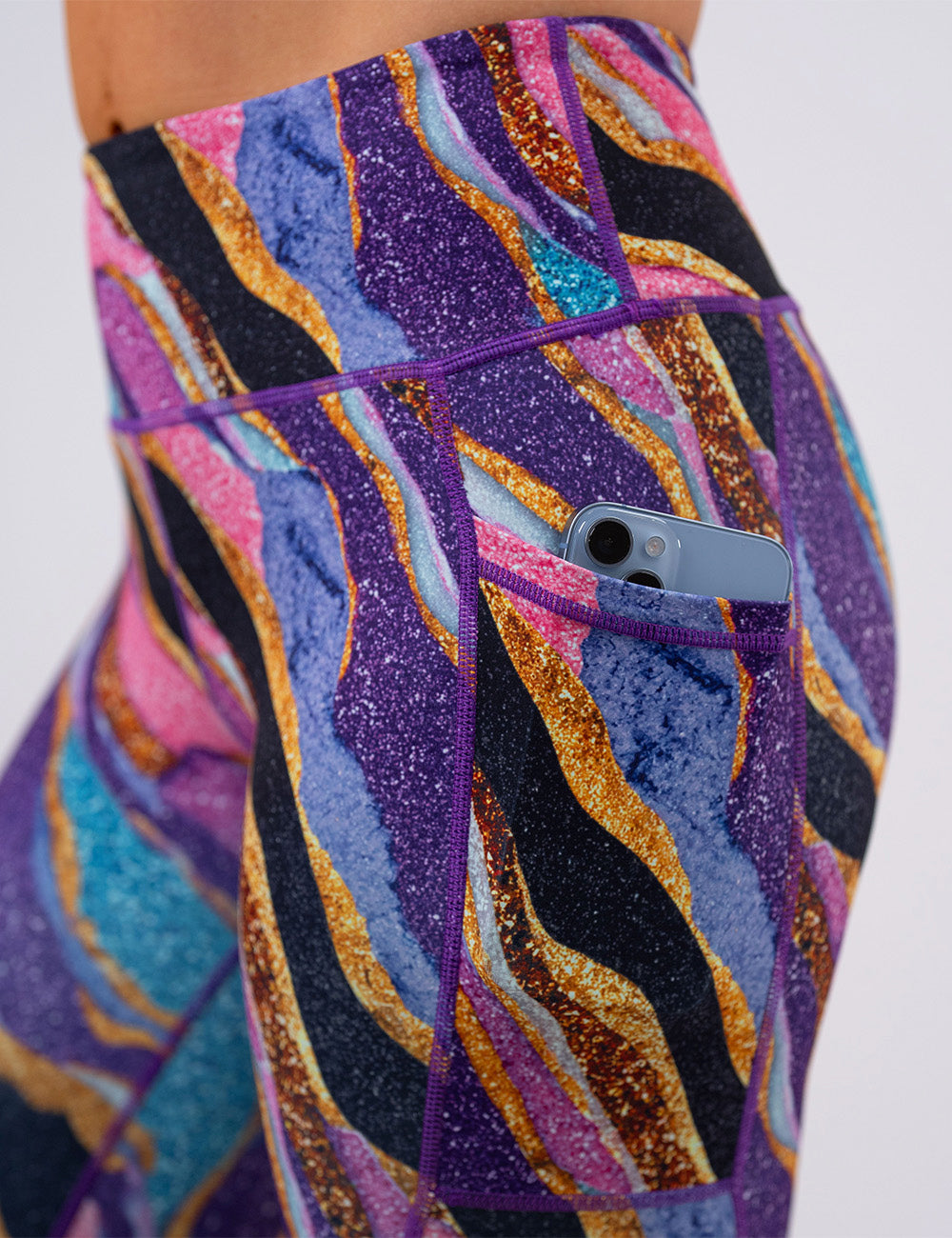 close up of the purple, gold, black, blue, and pink abstract stripes leggings with a phone in its pocket