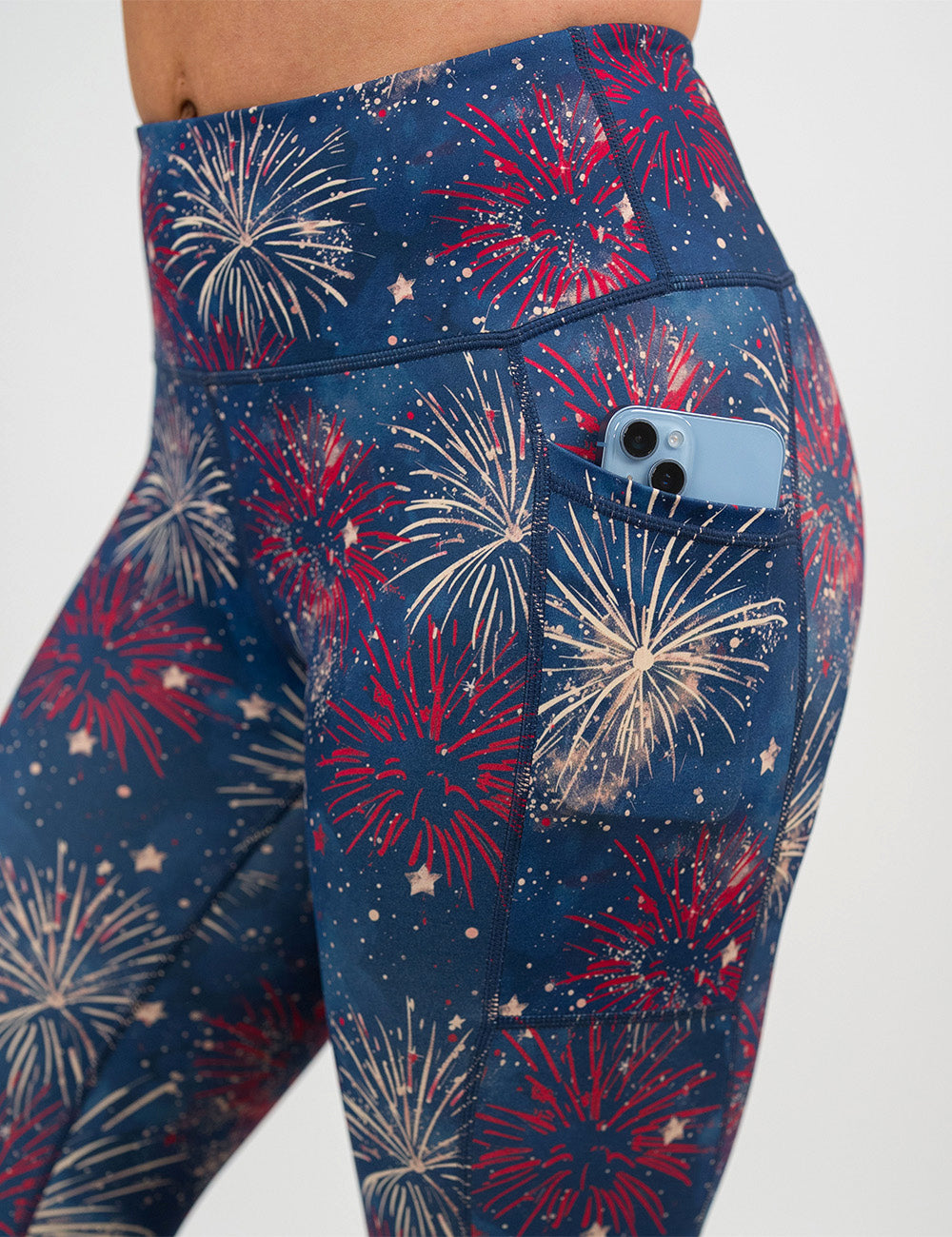 close up of the blue leggings with a red and white fireworks patterns pocket holding a phone against a white background