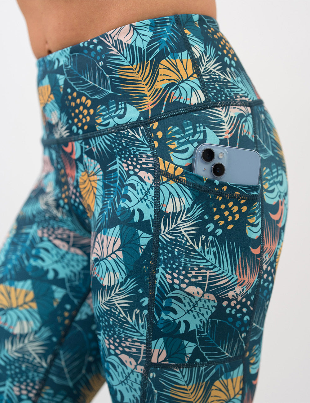 close up of a phone in the pocket of the blue, pink, yellow, and red tropical leaves leggings against a white background