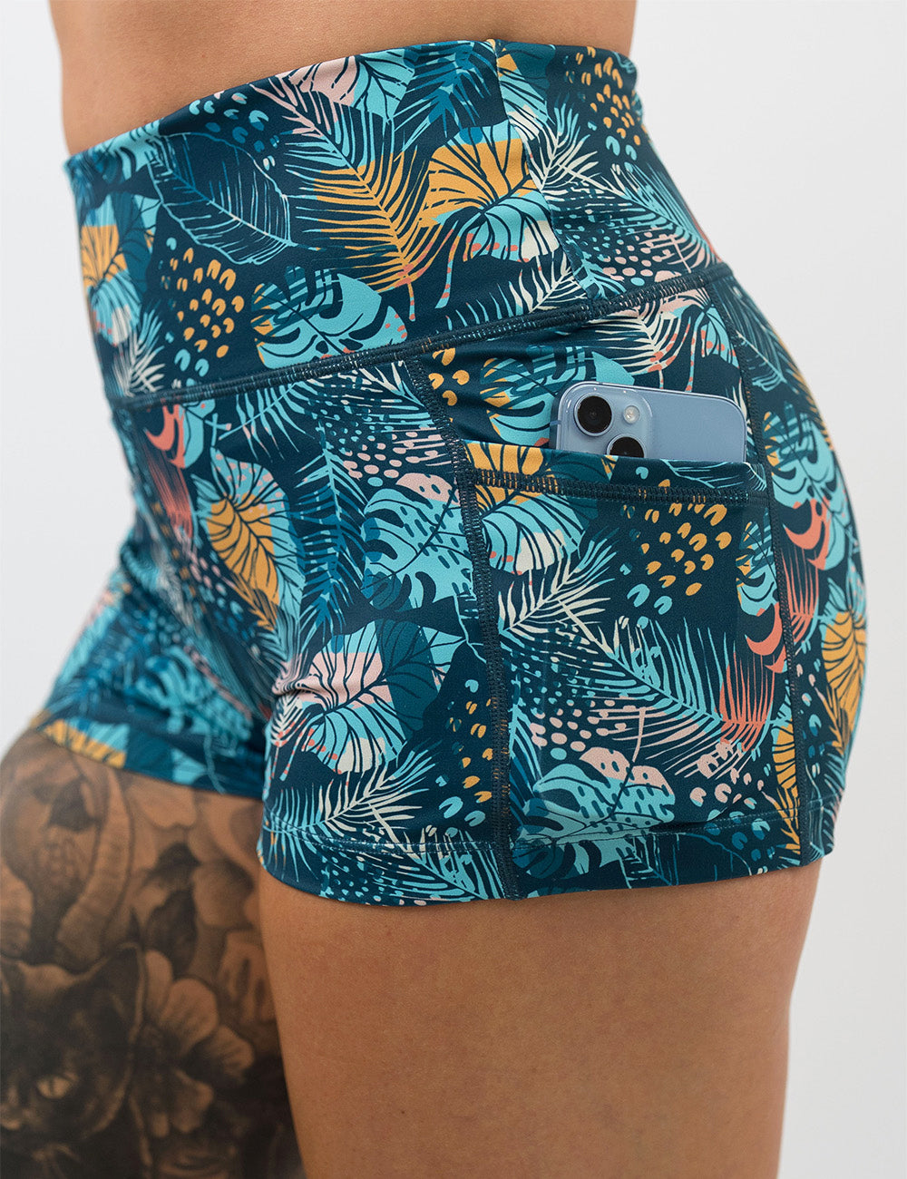 close up of a phone in the pocket of the 2.5 inch blue, pink, yellow, and red tropical leaves shorts against a white background
