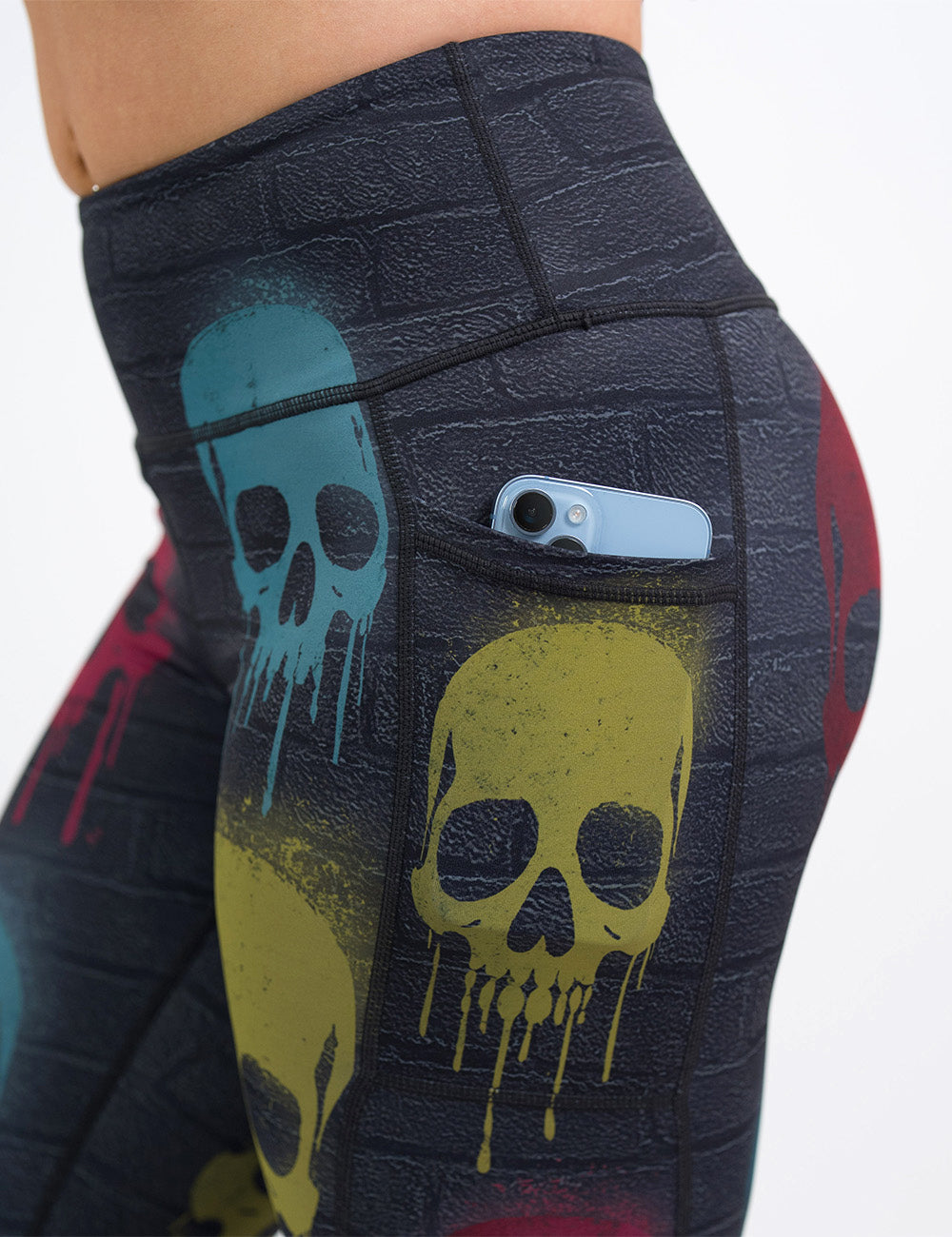 close up of the black brick leggings with spray painted red, yellow, and blue skulls all over with a phone in its pocket