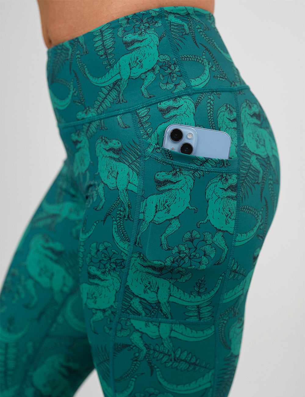 close up of a phone in the pocket of the teal t-rex pattern leggings against a white background