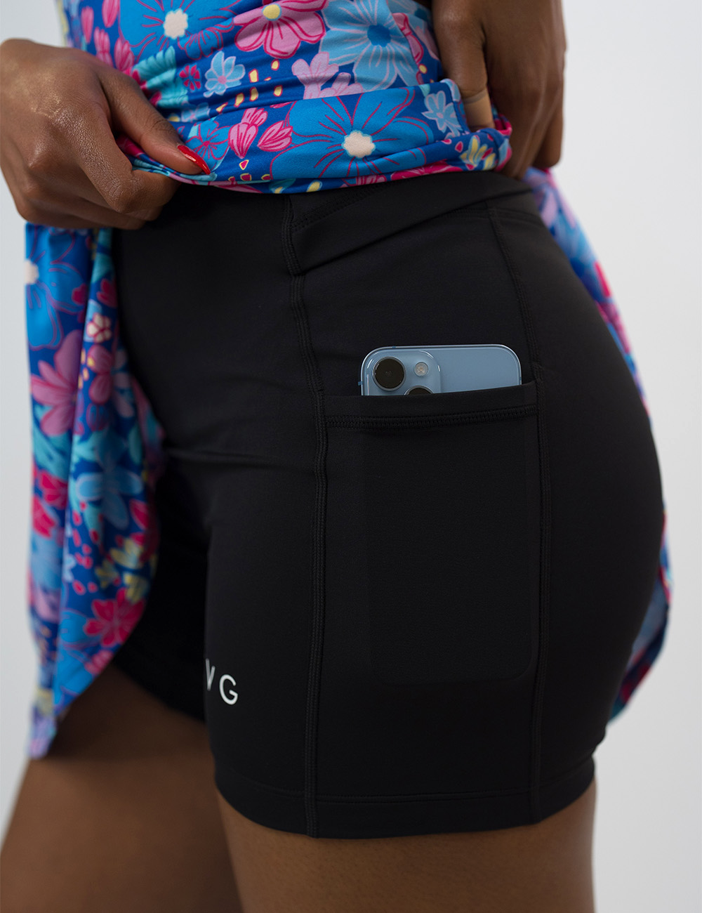 the colorful flowers active dress lifted up to show the built in shorts holding a phone in it pocket against a white background