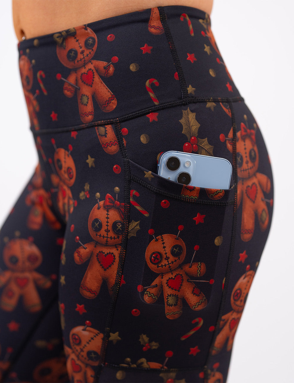 close up of the gingerbread voodoo dolls with pins in them surrounded by berries and stars on a black background leggings with a phone in its pocket