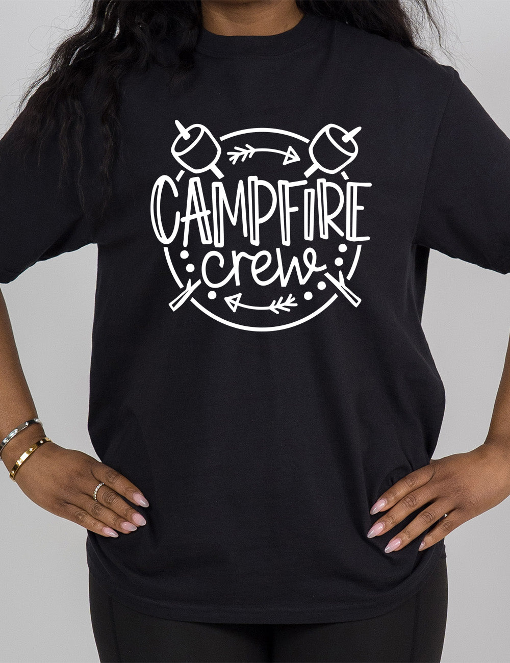 black modern tee with the words "campfire crew" in the middle of a circle, with a couple arrows and dots, and two marshmallows on sticks crossing behind the words