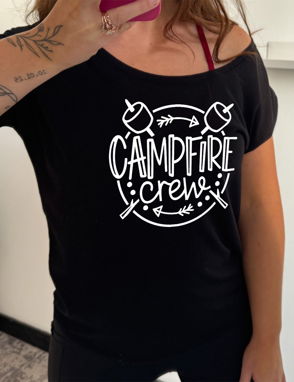 black slouchy tee with the words "campfire crew" in the middle of a circle, with a couple arrows and dots, and two marshmallows on sticks crossing behind the words